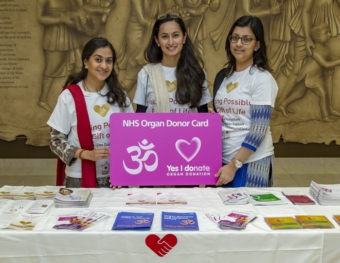 Three women stand behind a table of leaflets and hold an large organ donation card