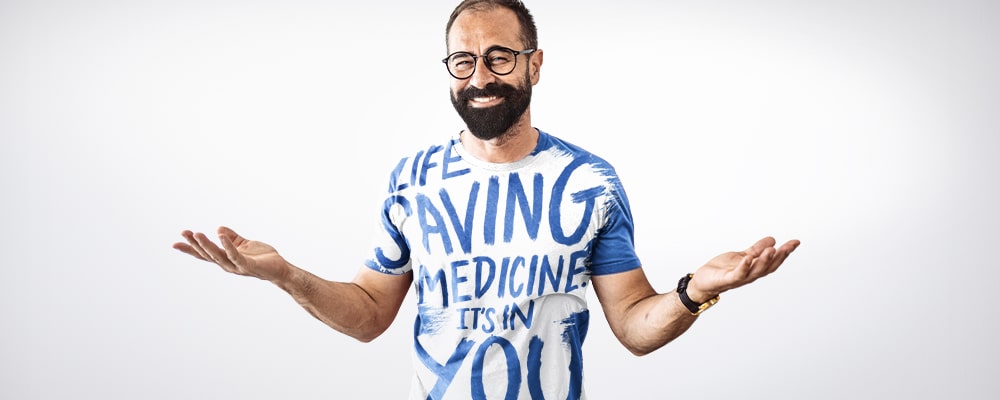 Man with a beard and glasses with his arms held up. He’s smiling and his t-shirt says Life saving medicine. It’s in you
