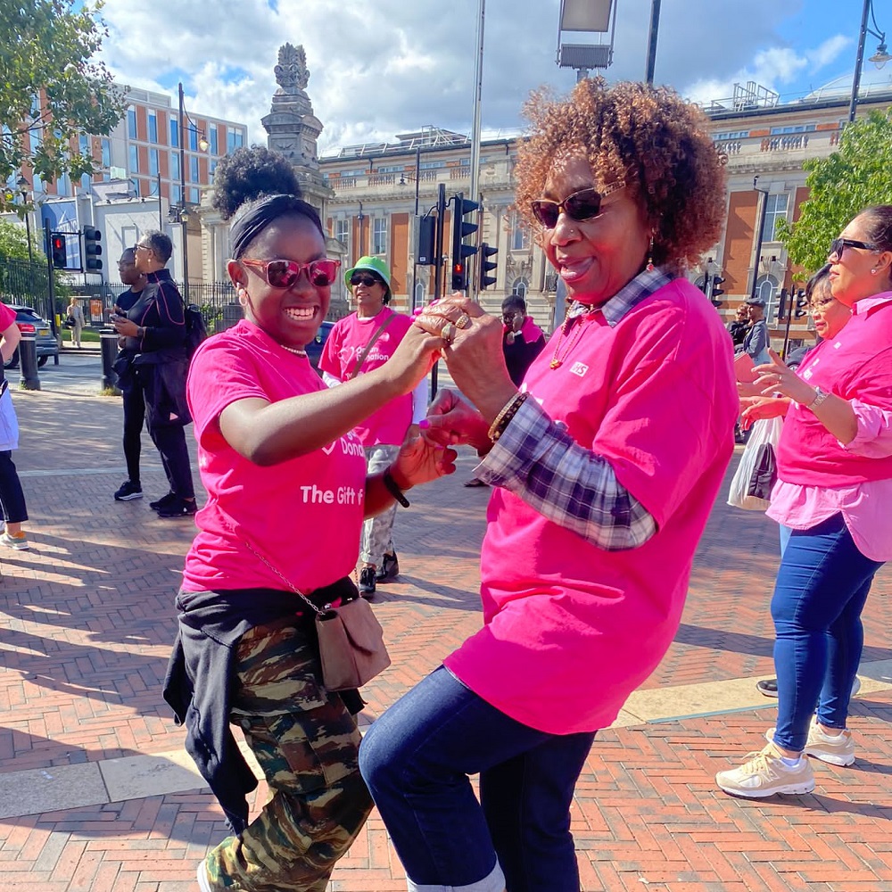 Two people dance at organ donation-themed flash mob event on a sunny day in Windrush Square, Brixton
