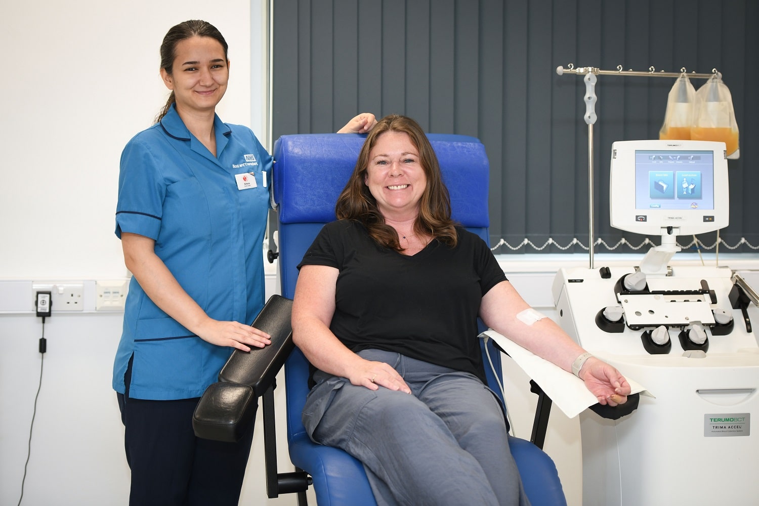 Platelet donor donating in chair with donor carer standing next to her smiling