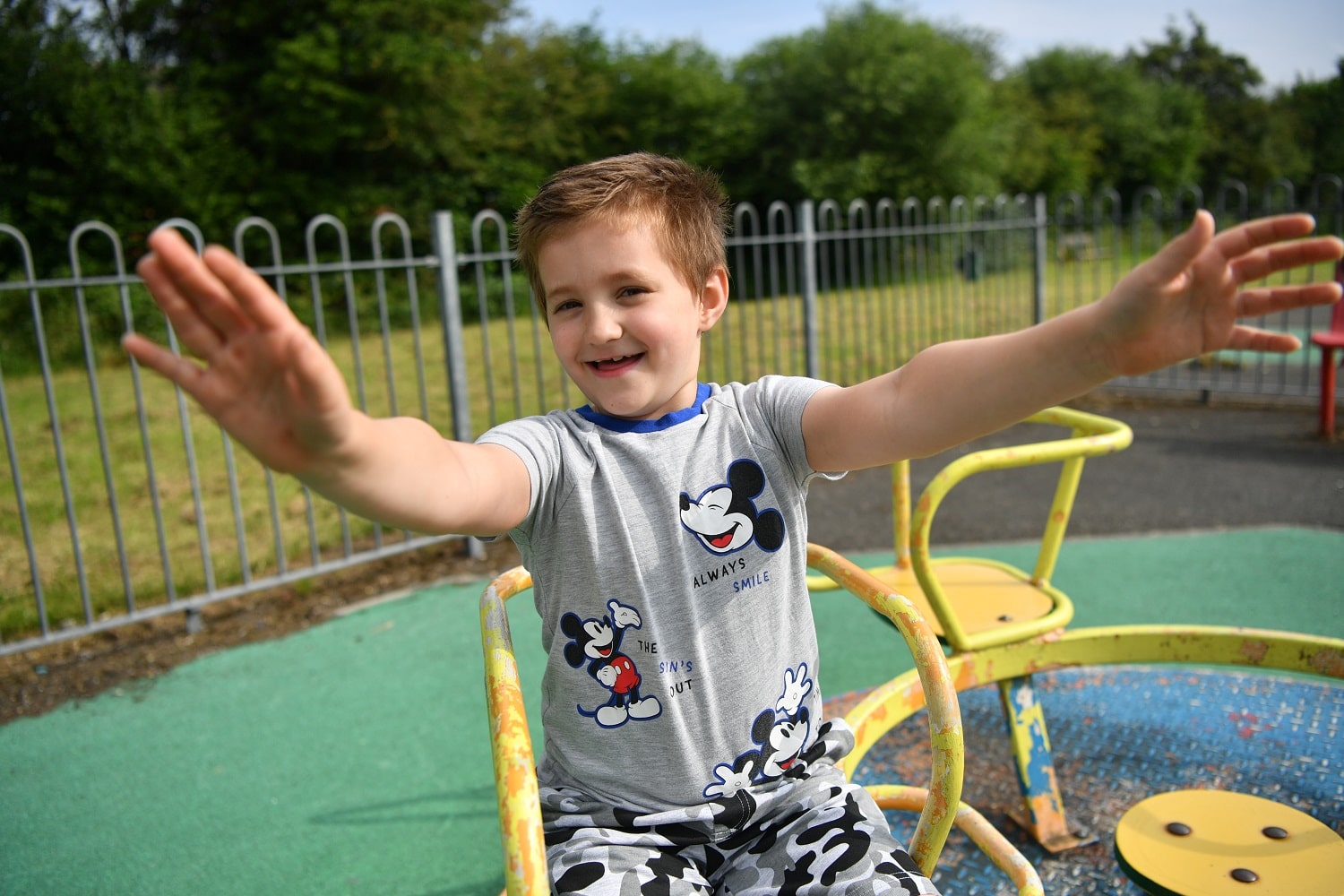 Harley sits on a playground roundabout smiling and holding arms in the air