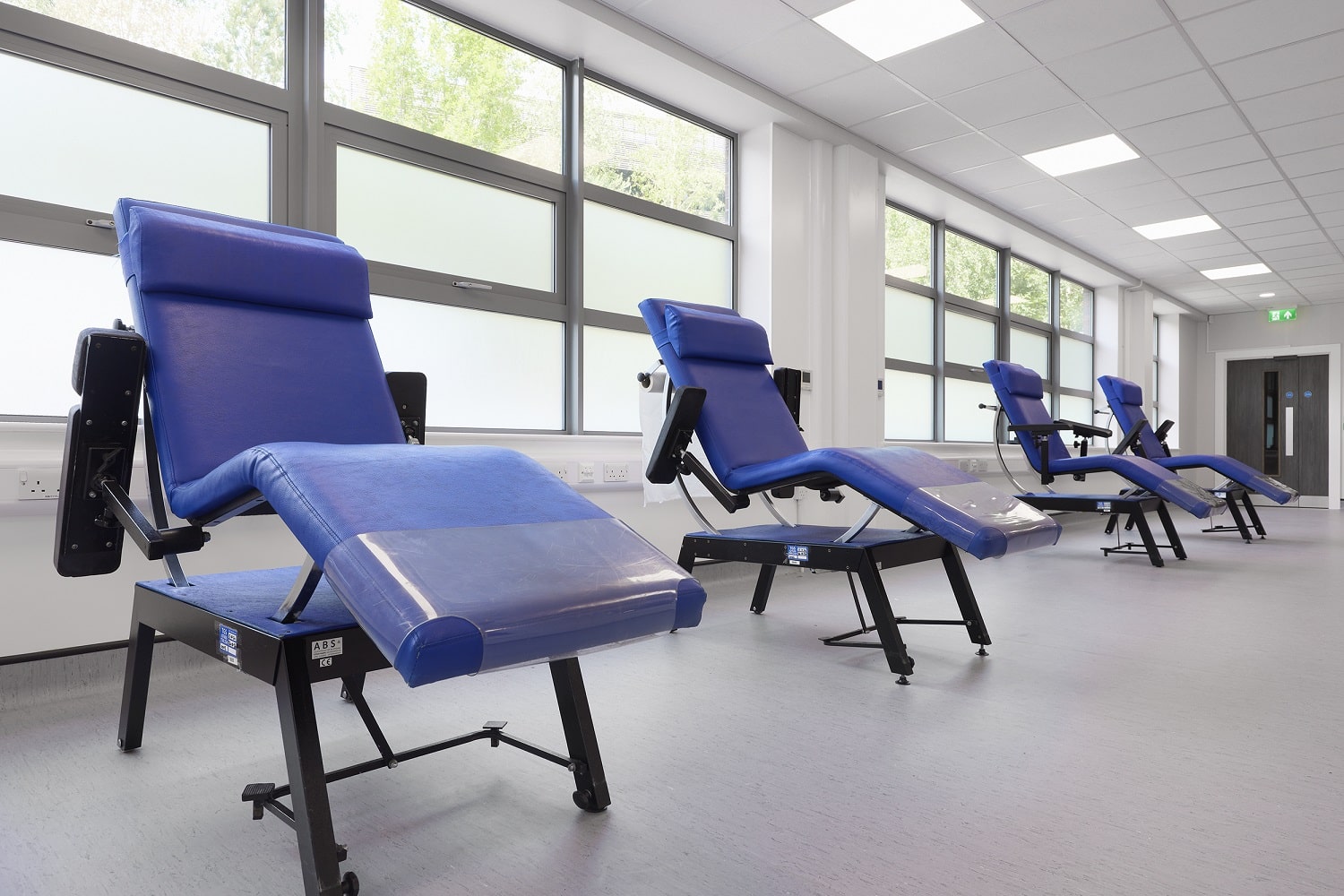 Four blood donation chairs in a row in a clean, clinical with lots of natural lighting from several windows behind the chairs