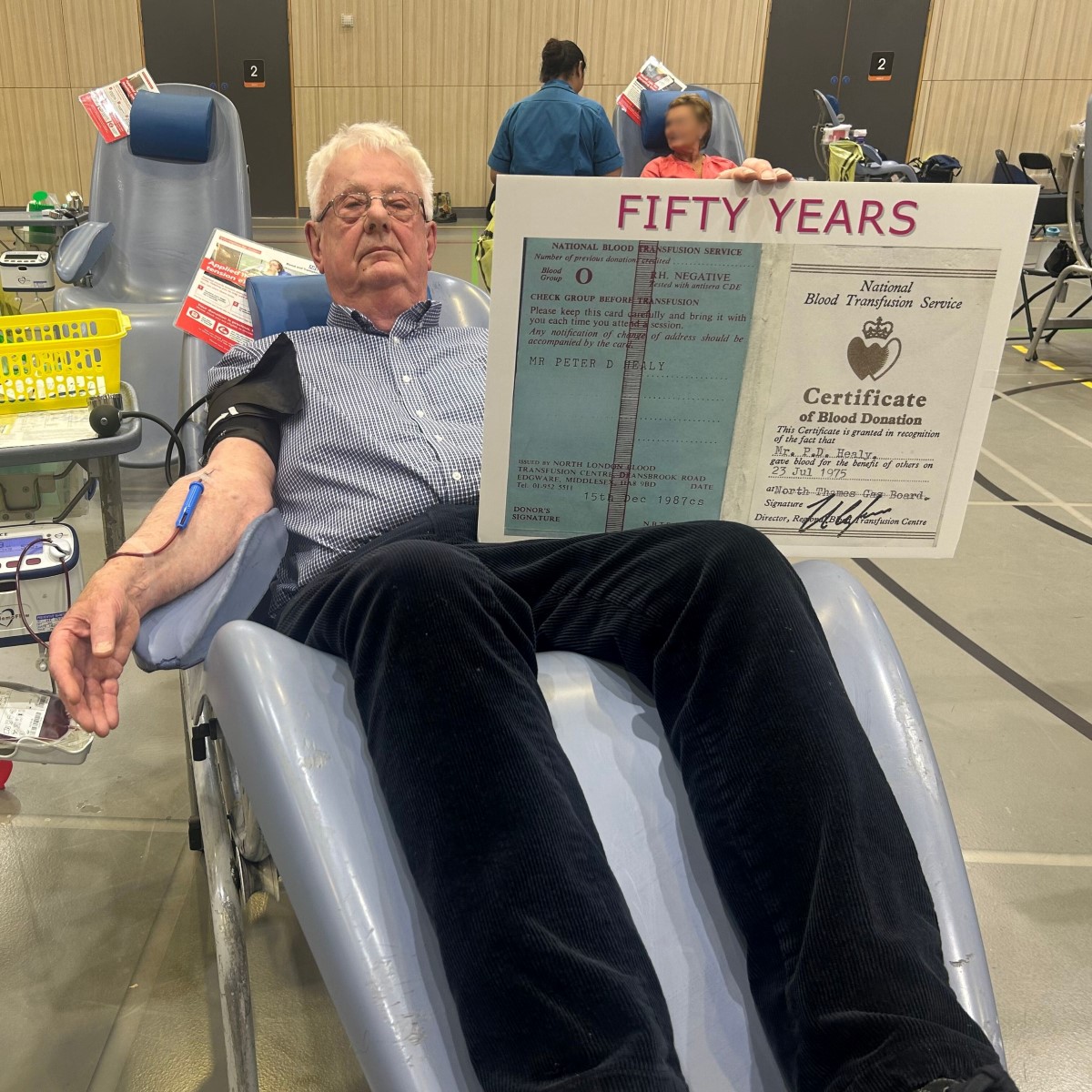 Pete Healy, sitting in a chair while donating blood. Holding a certificate showing his achievement of donating for 50 years.
