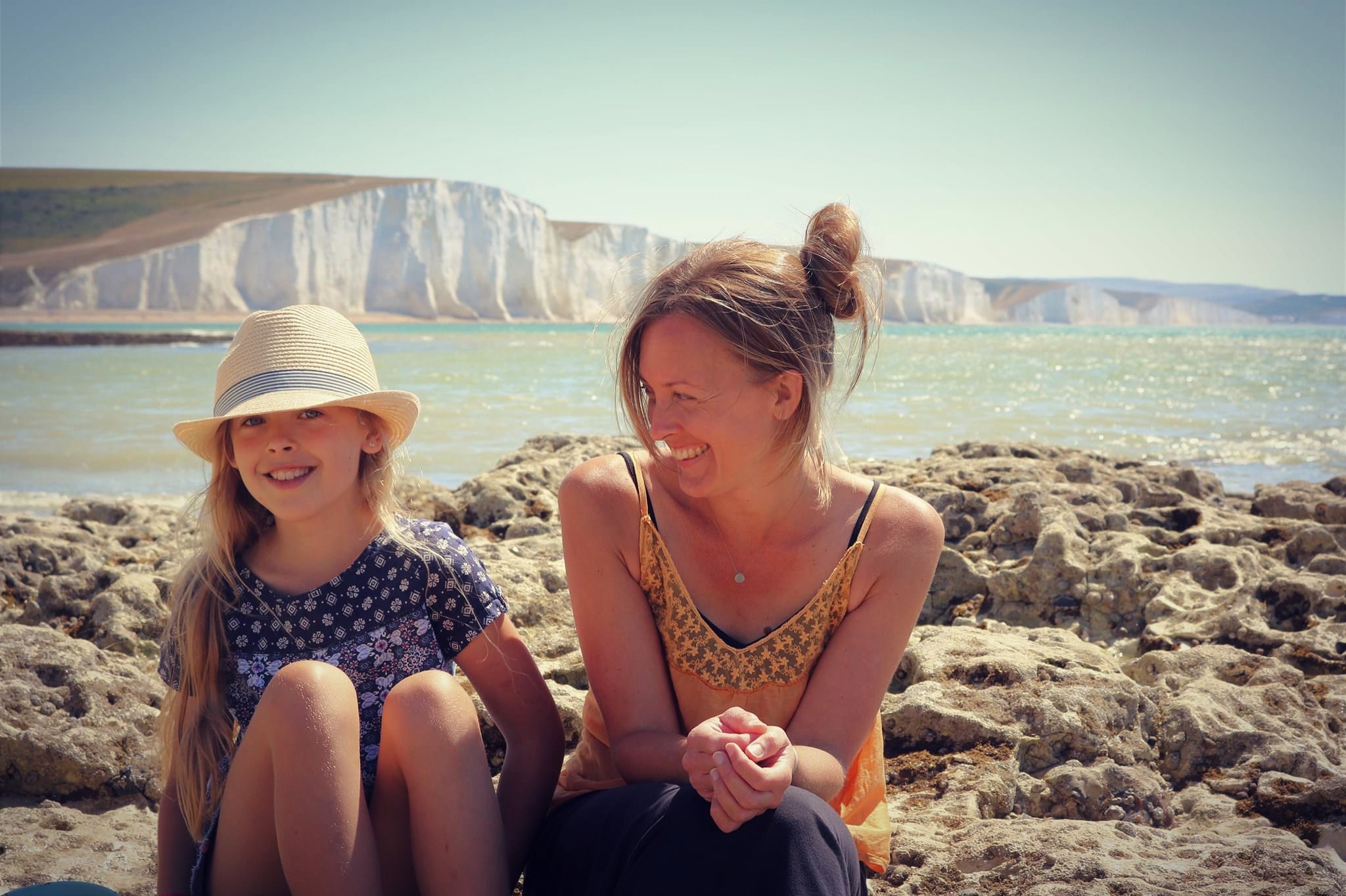 Amy and Issy sitting next to each other on the coast, Amy looking towards Issy with the sea behind them.