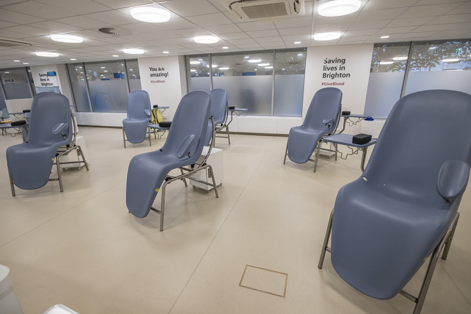 Donation area with several reclining chairs spaced apart, each with armrests and small side tables. Large windows line the back wall with posters reading messages like ‘You are amazing!’ and ‘Saving lives in Brighton'.