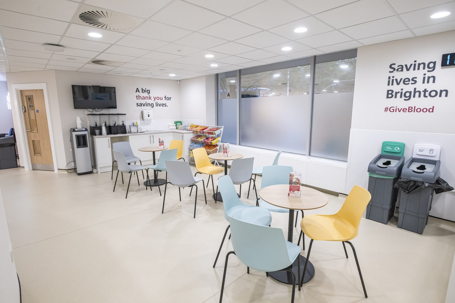 Refreshment area with round tables and colorful chairs, a counter with drinks and snacks, and bins for recycling. Wall signs read ‘A big thank you for saving lives’ and ‘Saving lives in Brighton #GiveBlood.’.
