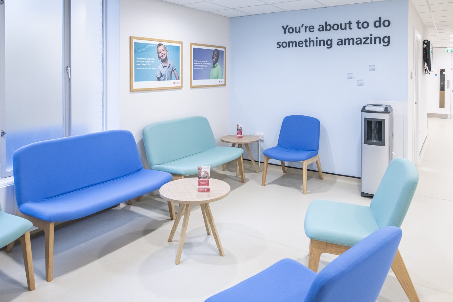 Bright waiting area with blue and teal chairs around small wooden tables, posters on the wall, and text reading ‘You’re about to do something amazing.’ A water dispenser is in the corner.