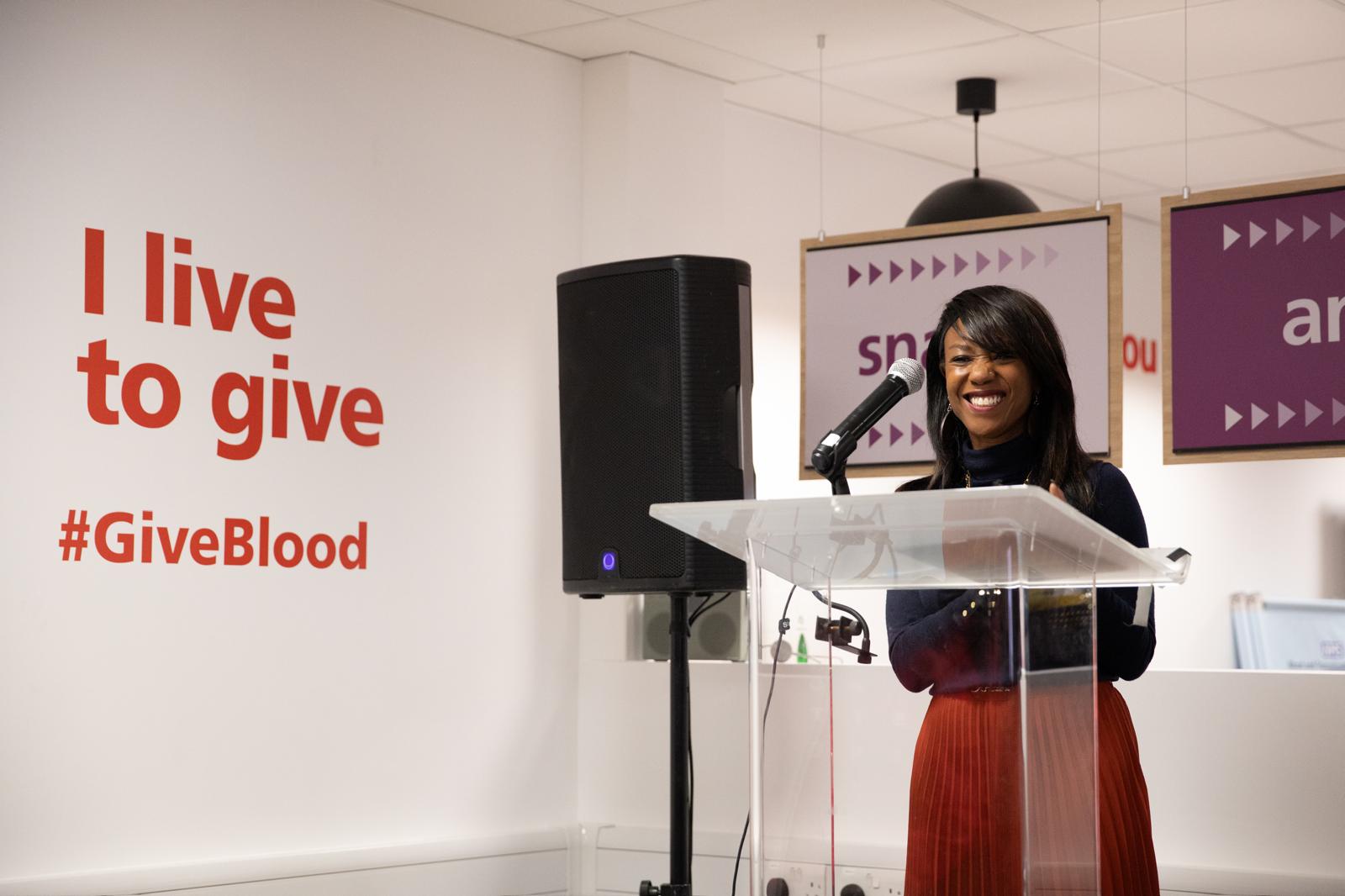Dr Debbie Weekes-Bernard standing at a podium, next to wall text stating "I live to give #GiveBlood"