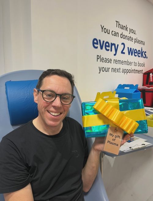 A donor, smiling and holding up a present labelled 'My gift of Plasma'