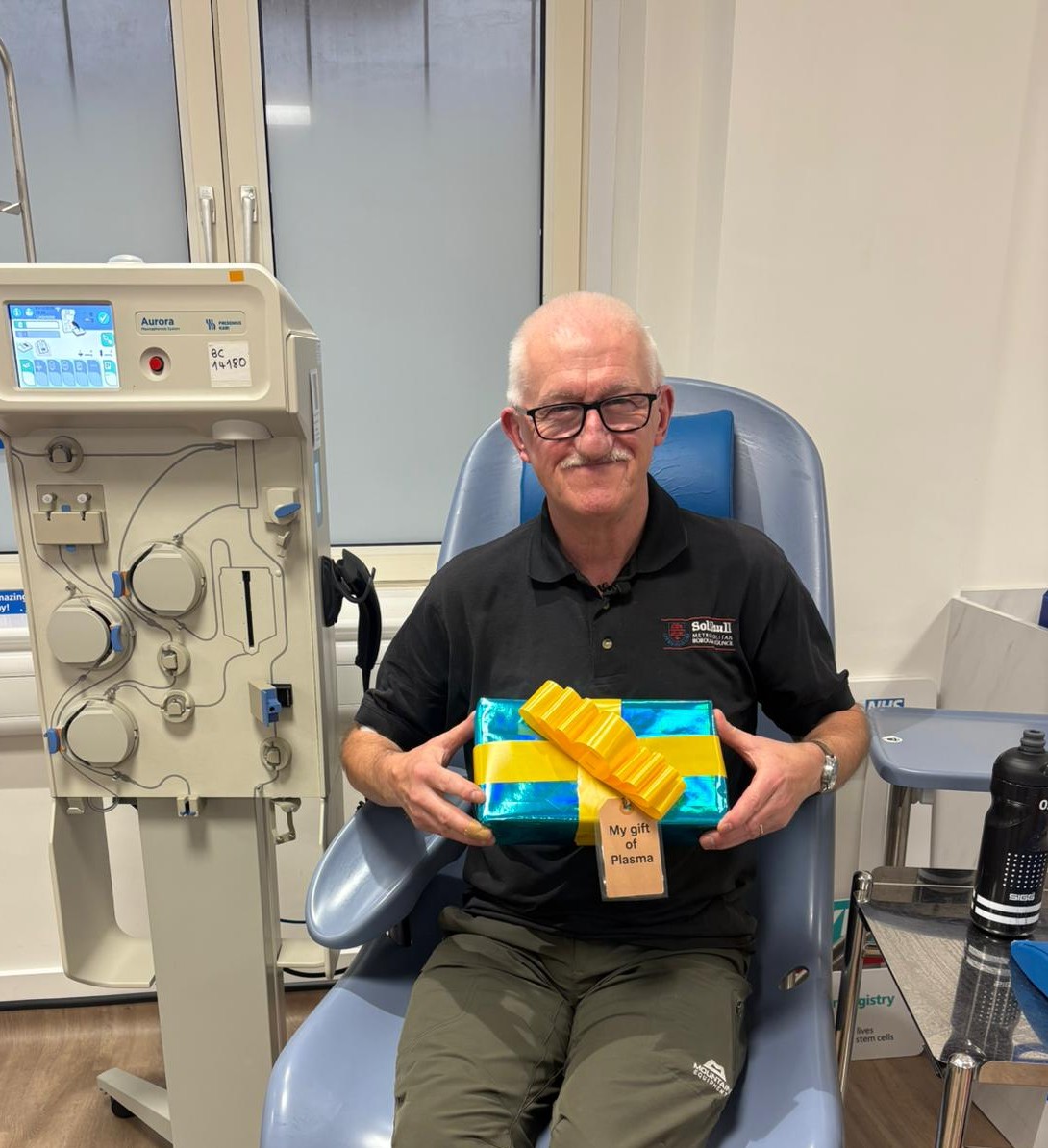 Andy Stowe, sitting in a donation chair and holding a present box. The box is wrapped in light blue wrapping with a yellow bow and has the tag "my gift of plasma"