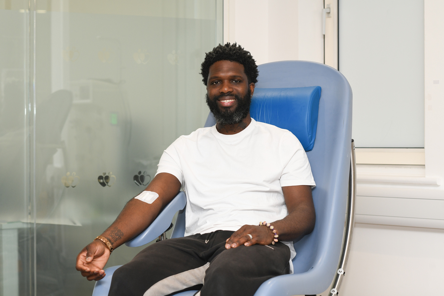 A man of Black heritage smiles in donation chair after just donated, a plaster on his arm.