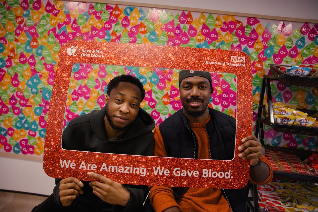 Two donors who booked as a group look through a 'selfie frame' that say 'We Are Amazing, We Gave Blood.'